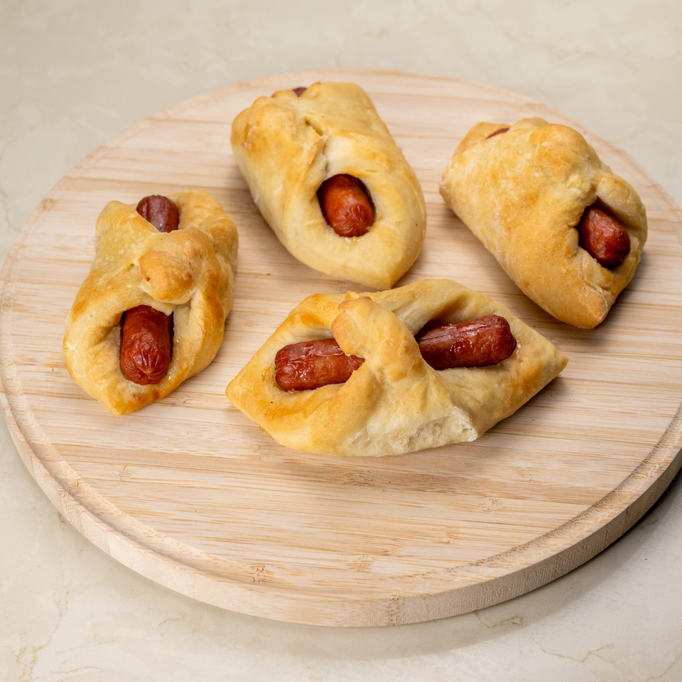 Four golden-brown Manske rolls, each with a hot dog peeking out, on a wooden cutting board.