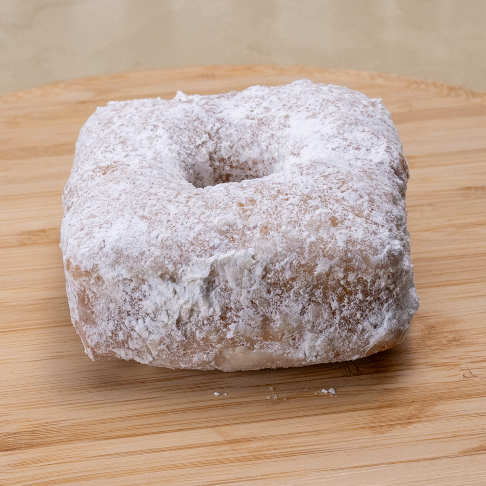 A square Manske Roll donut, covered in powdered sugar, rests on a light wooden surface.
